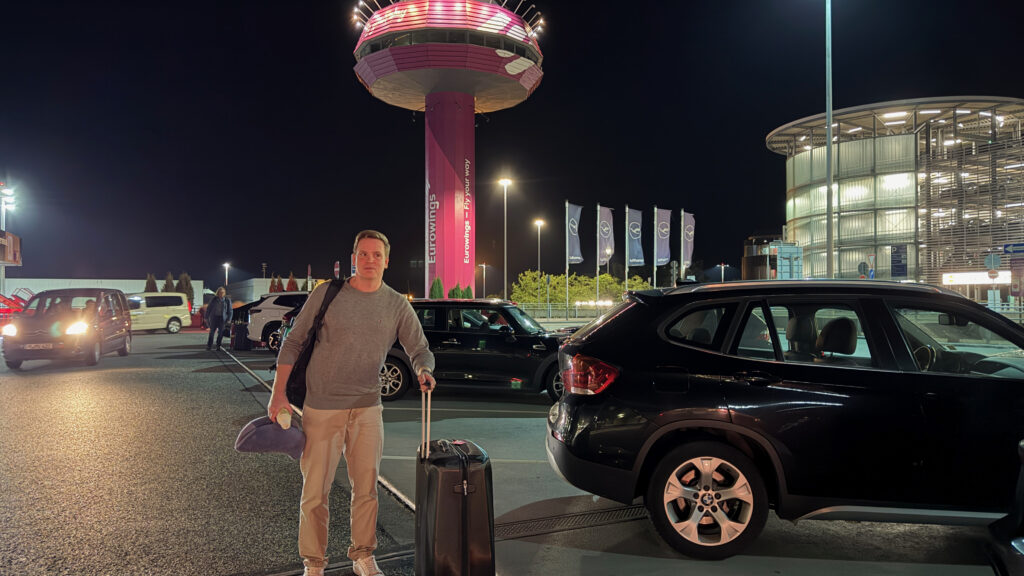 Sascha mit Koffer vor dem Flughafen Hannover und pink beleuchtetem Turm